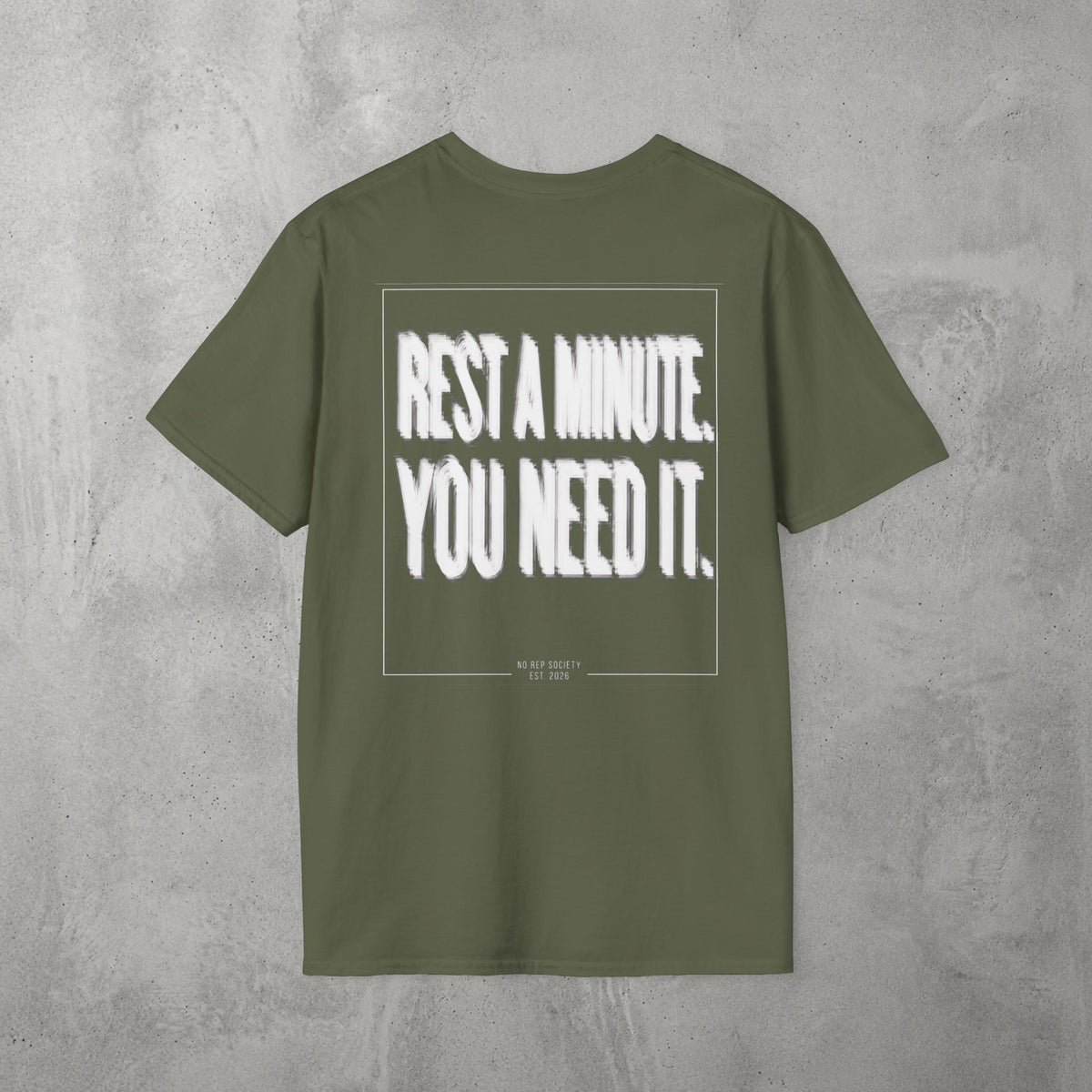 Military Green Rest A Minute Tee by The No Rep Society, flat lay on grey concrete, back print of REST A MINUTE. YOU NEED IT. in bold blurred capitals inside a thin frame.
