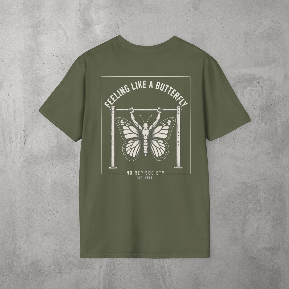 Military Green Feeling Like A Butterfly Tee by The No Rep Society, flat lay on grey concrete, back print of a moth with spread wings hanging between two rig uprights under FEELING LIKE A BUTTERFLY lettering.