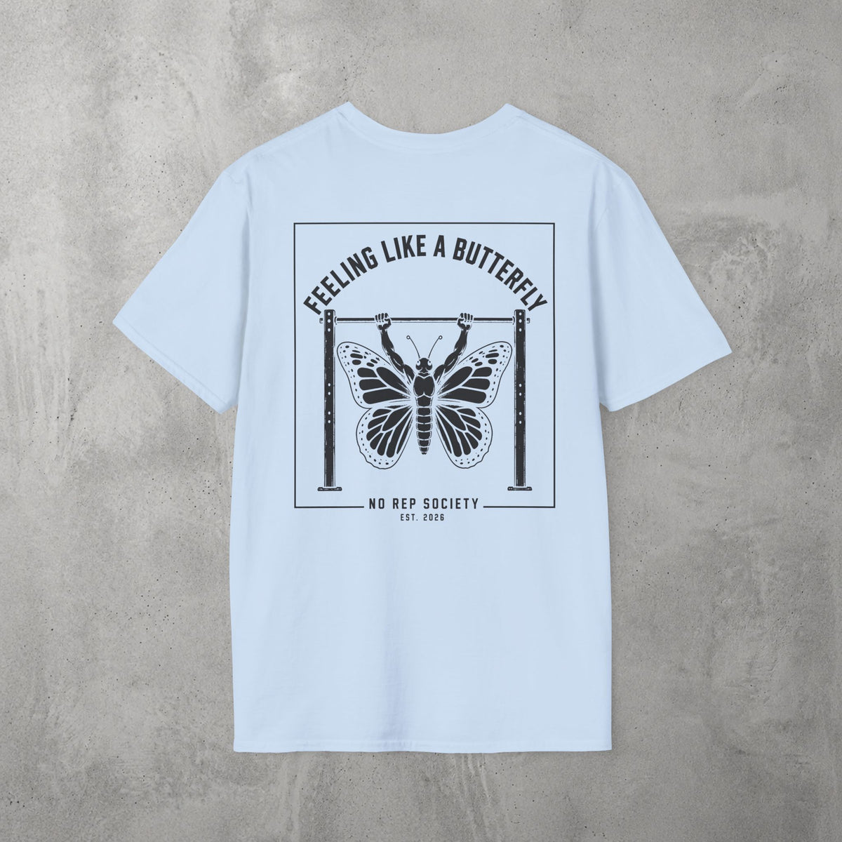 Light Blue Feeling Like A Butterfly Tee by The No Rep Society, flat lay on grey concrete, back print of a moth with spread wings hanging between two rig uprights under FEELING LIKE A BUTTERFLY lettering.