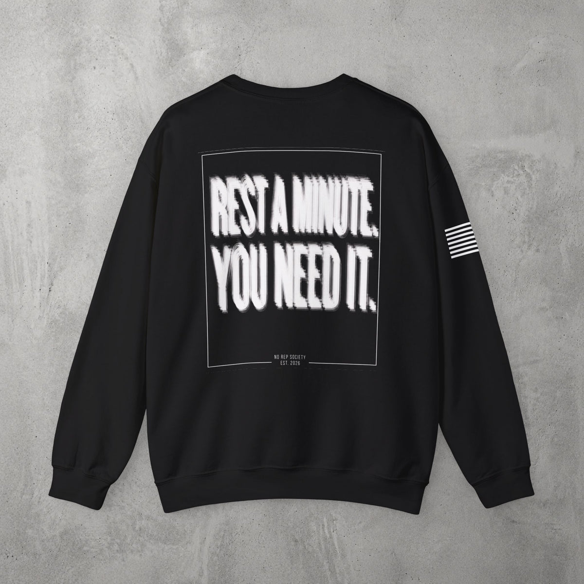 Black Rest A Minute Crewneck by The No Rep Society, flat lay on grey concrete, back print of REST A MINUTE. YOU NEED IT. in bold blurred capitals inside a thin frame.