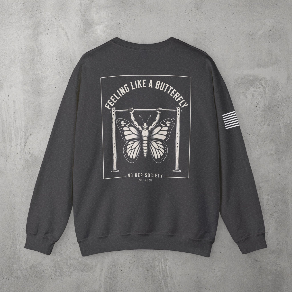 Dark Heather Feeling Like A Butterfly Crewneck by The No Rep Society, flat lay on grey concrete, back print of a moth with spread wings hanging between two rig uprights under FEELING LIKE A BUTTERFLY lettering.