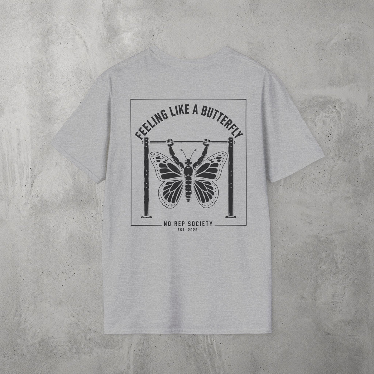 Sport Grey Feeling Like A Butterfly Tee by The No Rep Society, flat lay on grey concrete, back print of a moth with spread wings hanging between two rig uprights under FEELING LIKE A BUTTERFLY lettering.