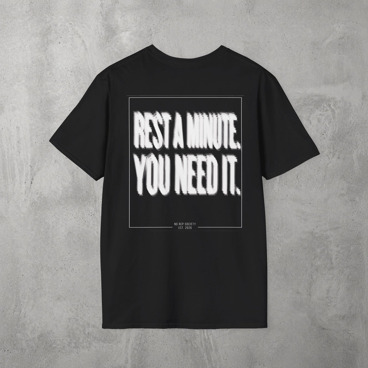 Black Rest A Minute Tee by The No Rep Society, flat lay on grey concrete, back print of REST A MINUTE. YOU NEED IT. in bold blurred capitals inside a thin frame.