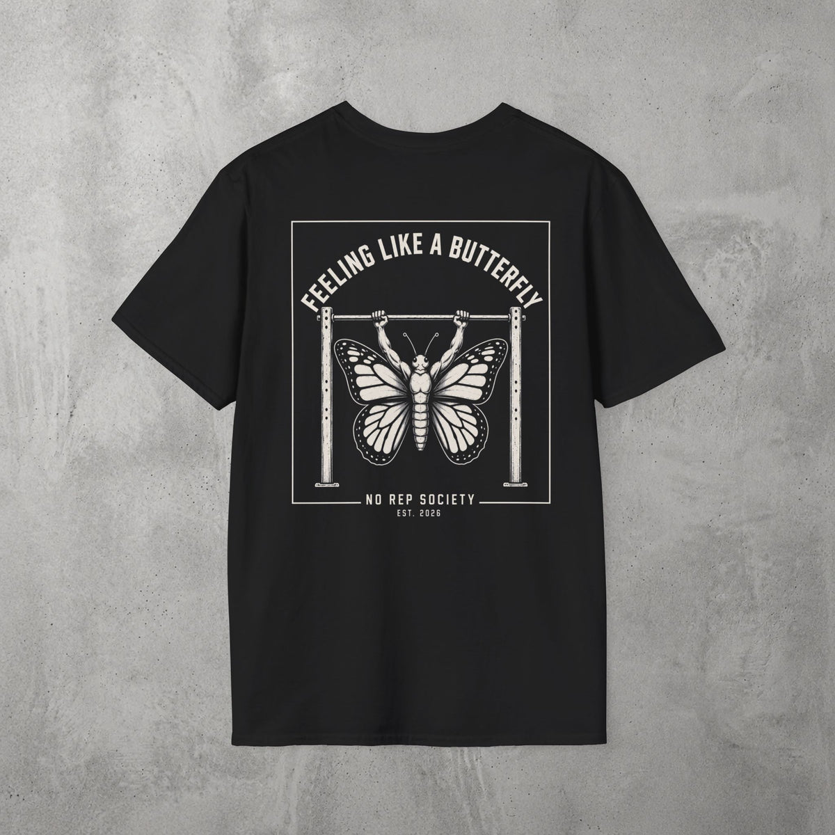 Black Feeling Like A Butterfly Tee by The No Rep Society, flat lay on grey concrete, back print of a moth with spread wings hanging between two rig uprights under FEELING LIKE A BUTTERFLY lettering.