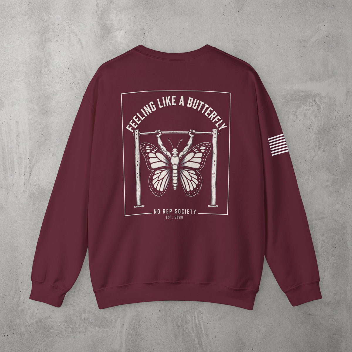 Maroon Feeling Like A Butterfly Crewneck by The No Rep Society, flat lay on grey concrete, back print of a moth with spread wings hanging between two rig uprights under FEELING LIKE A BUTTERFLY lettering.