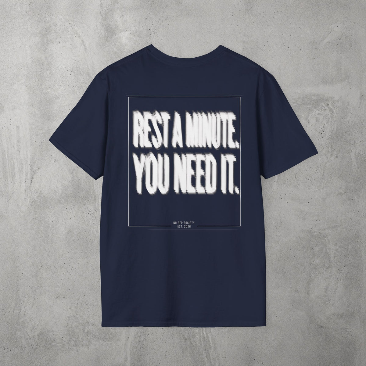 Navy Rest A Minute Tee by The No Rep Society, flat lay on grey concrete, back print of REST A MINUTE. YOU NEED IT. in bold blurred capitals inside a thin frame.