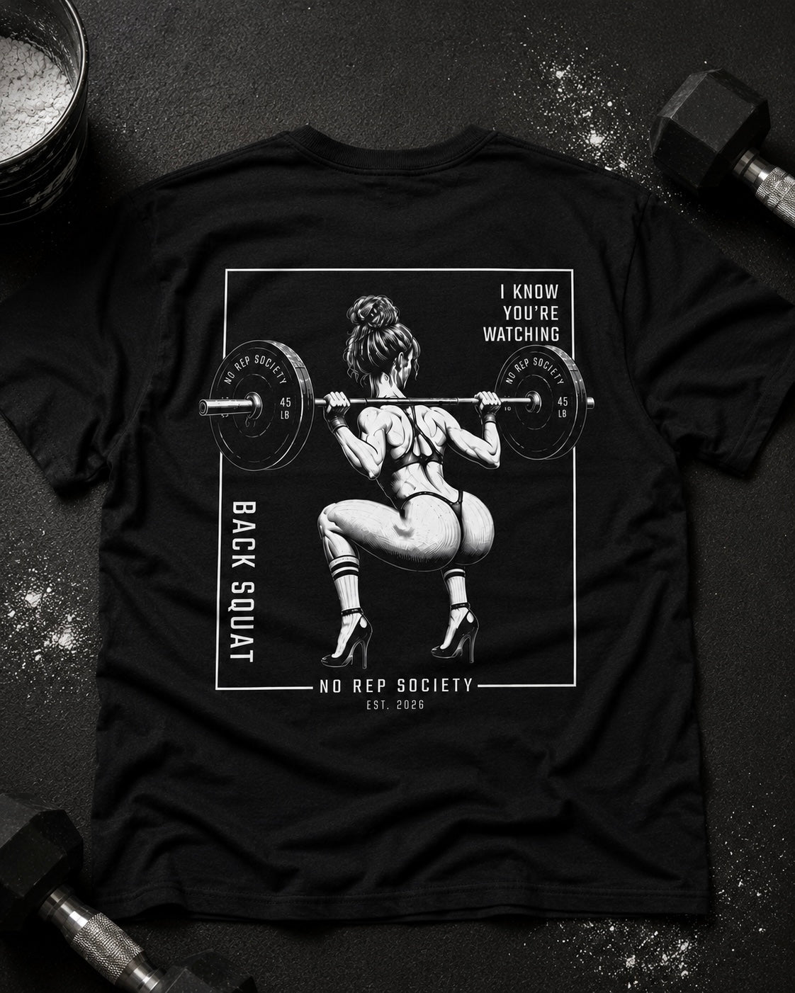 Black I Know You're Watching Tee by The No Rep Society, laid flat on a dark gym floor between a chalk bucket and dumbbells, back print of a woman mid back-squat inside a bordered frame under I KNOW YOU'RE WATCHING lettering.