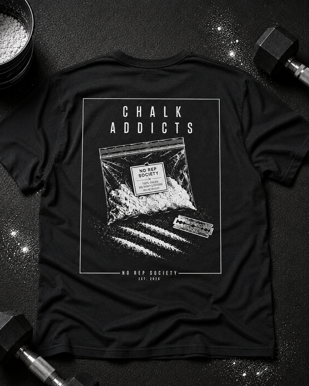 Black Chalk Addicts Tee by The No Rep Society, laid flat on a dark gym floor between a chalk bucket and a dumbbell, back print of an open bag of gym chalk spilling across a bordered frame under CHALK ADDICTS lettering.