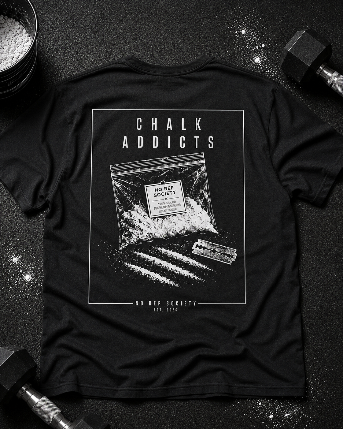 Black Chalk Addicts Tee by The No Rep Society, laid flat on a dark gym floor between a chalk bucket and a dumbbell, back print of an open bag of gym chalk spilling across a bordered frame under CHALK ADDICTS lettering.