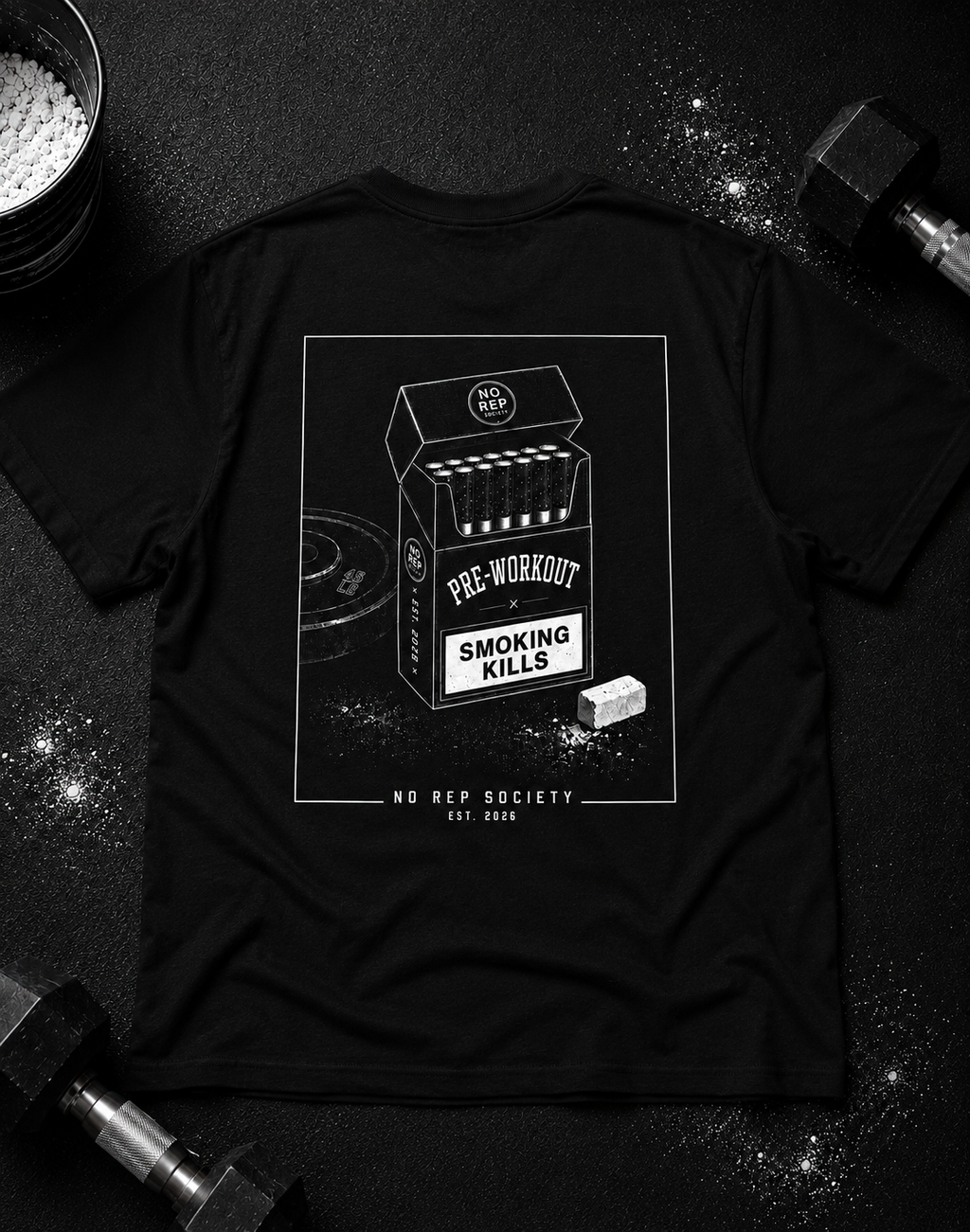 Black Pre-Workout Tee by The No Rep Society, laid flat on a dark gym floor between a chalk bucket and dumbbells, back print of an open cigarette pack labelled PRE-WORKOUT with a SMOKING KILLS warning and a scoop spilling powder.