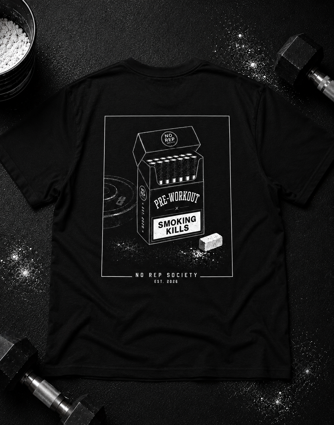 Black Pre-Workout Tee by The No Rep Society, laid flat on a dark gym floor between a chalk bucket and dumbbells, back print of an open cigarette pack labelled PRE-WORKOUT with a SMOKING KILLS warning and a scoop spilling powder.