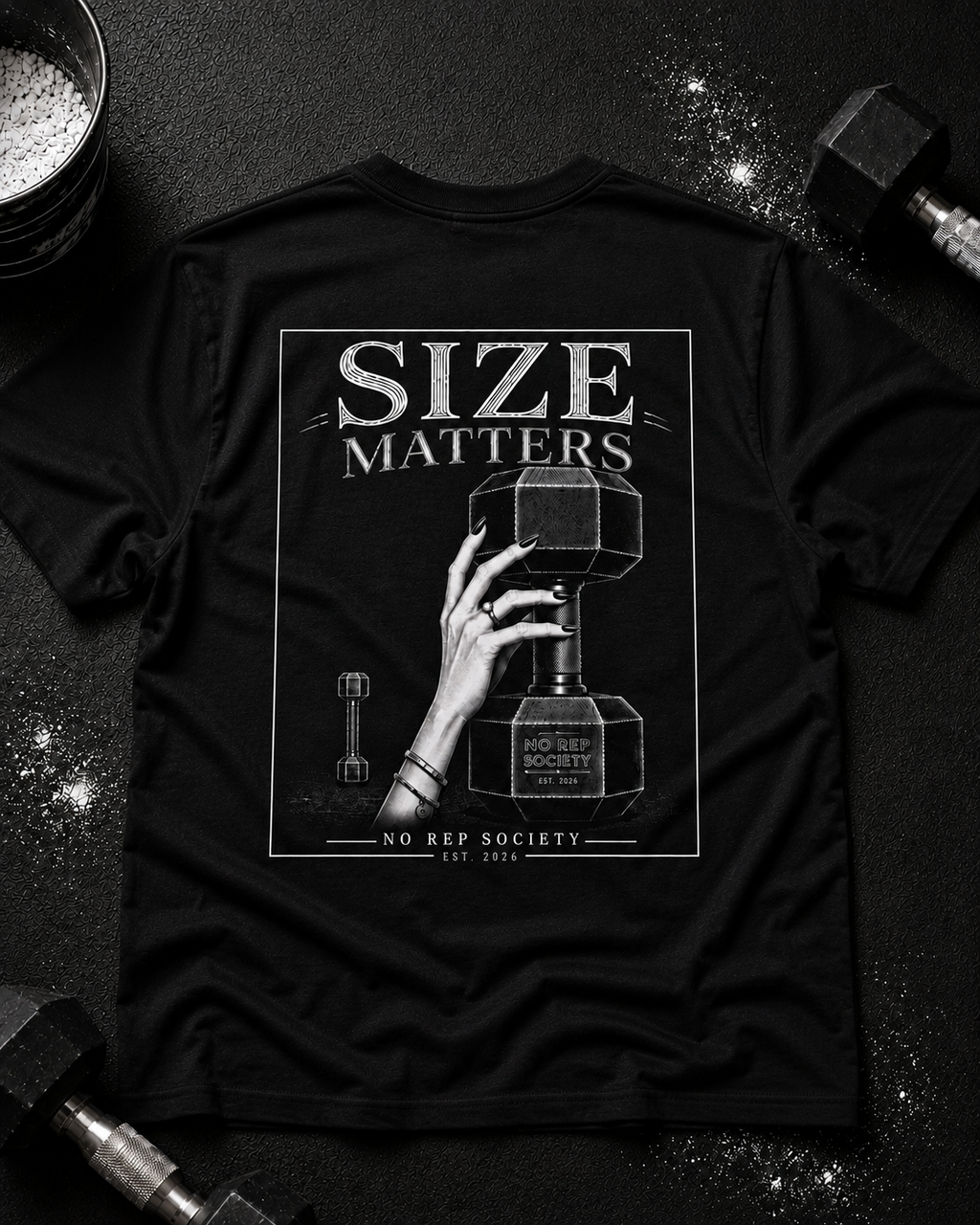 Black Size Matters Tee by The No Rep Society, laid flat on a dark gym floor between a chalk bucket and a dumbbell, back print of a hand gripping a large weight plate under SIZE MATTERS lettering.
