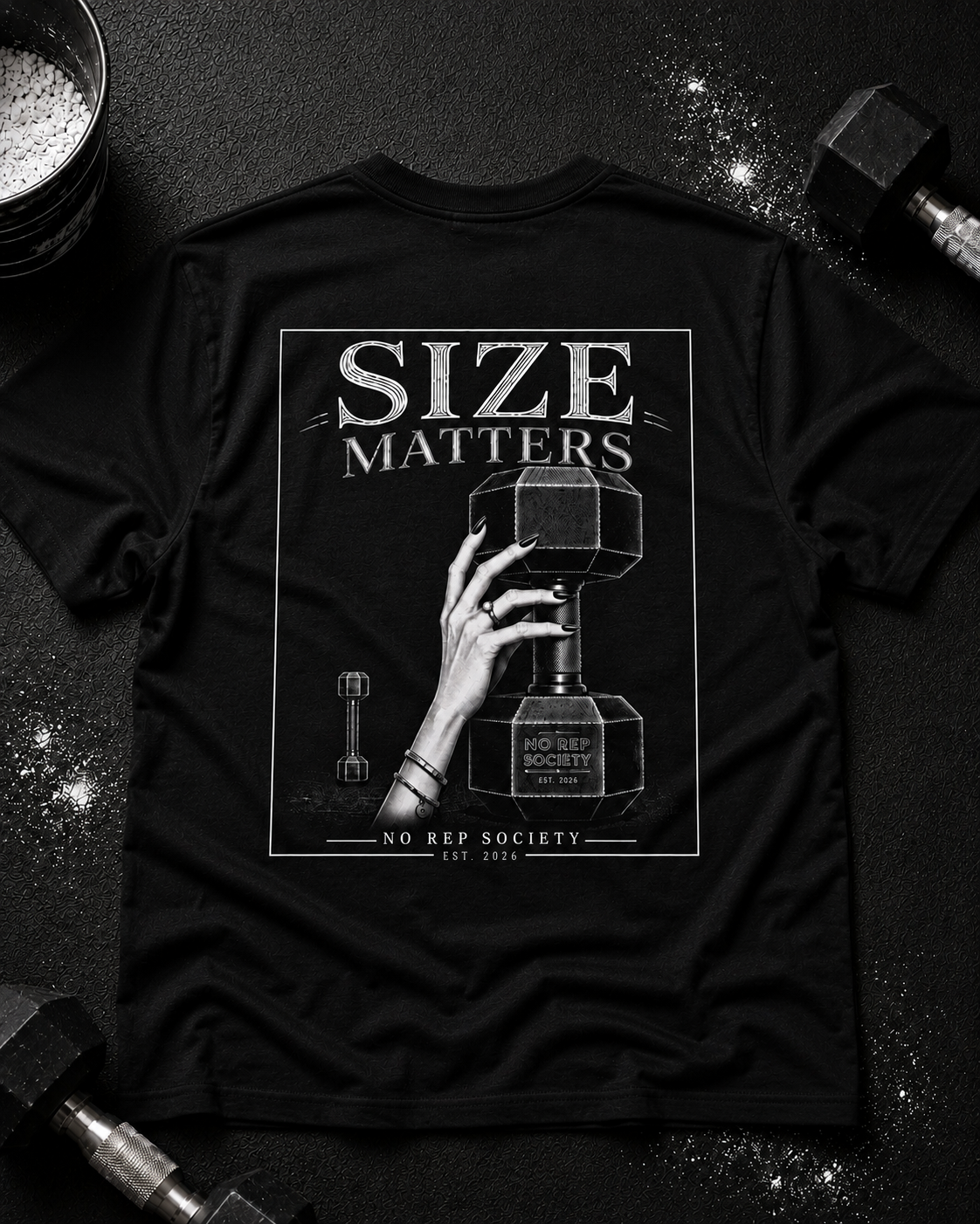Black Size Matters Tee by The No Rep Society, laid flat on a dark gym floor between a chalk bucket and a dumbbell, back print of a hand gripping a large weight plate under SIZE MATTERS lettering.